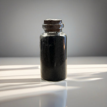 Small glass bottle with cork lid containing a black salt, on a reflective surface.